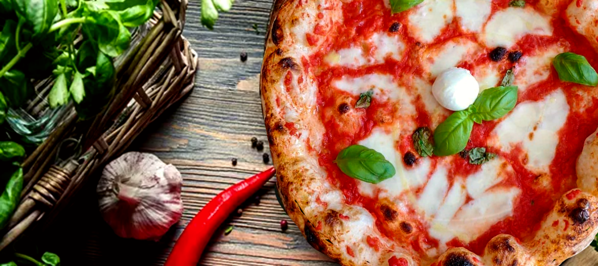 Margherita pizza on dark wood surrounded by fresh basil, garlic, and chili pepper